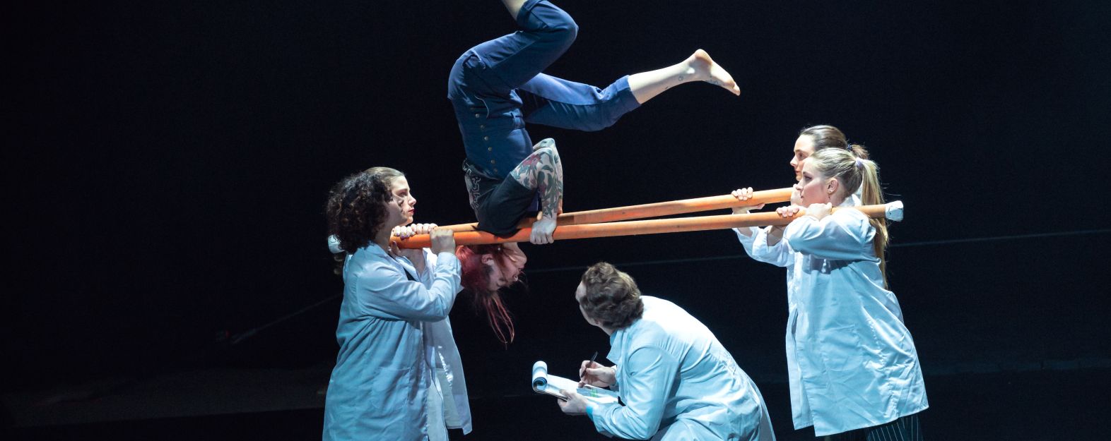 Several performers in lab coats holding up two parallel poles on which another performer is posed upside down