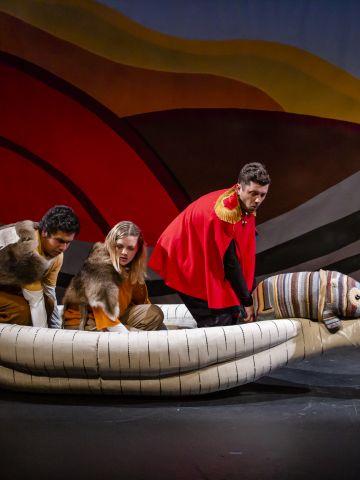 Three perfomers and a puppet wombat in a canoe on stage.The performers are looking into the water at one side of the canoe. Two performers are wearing animal skins