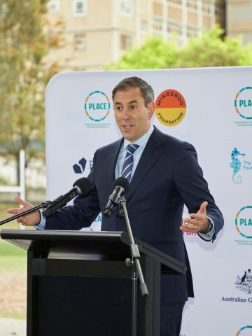 Treasurer Jim Clamers at a lectern with micrphones in front of a banner with logos of PLACE and government and philanthropic funders