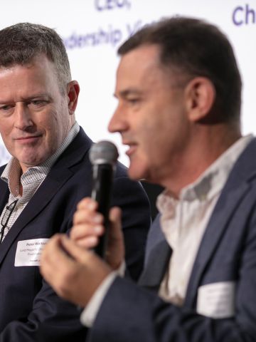 One woman and two men sitting next to each other on a panel discussion. One man is holdign a mcirophone and speaking