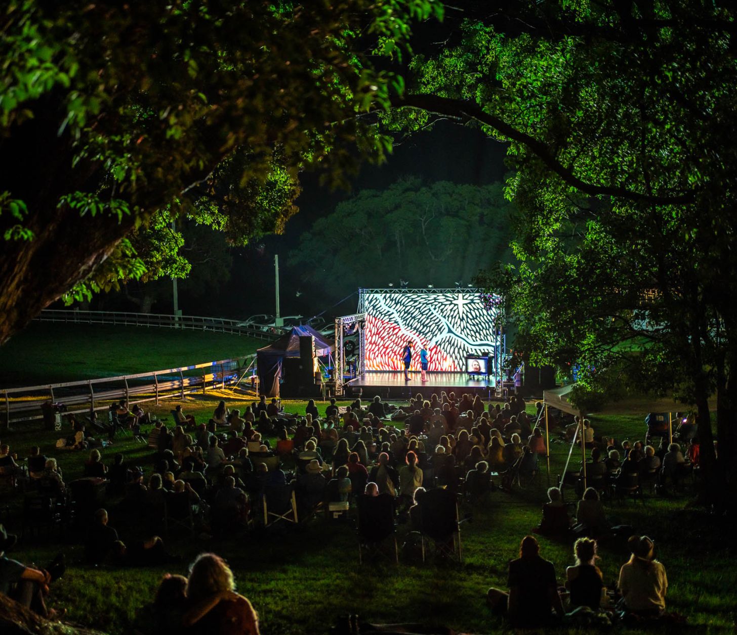 View of an outdoor night performance seen through a space in trees with the audience looking at a small stage with a projected image as the backdrop.