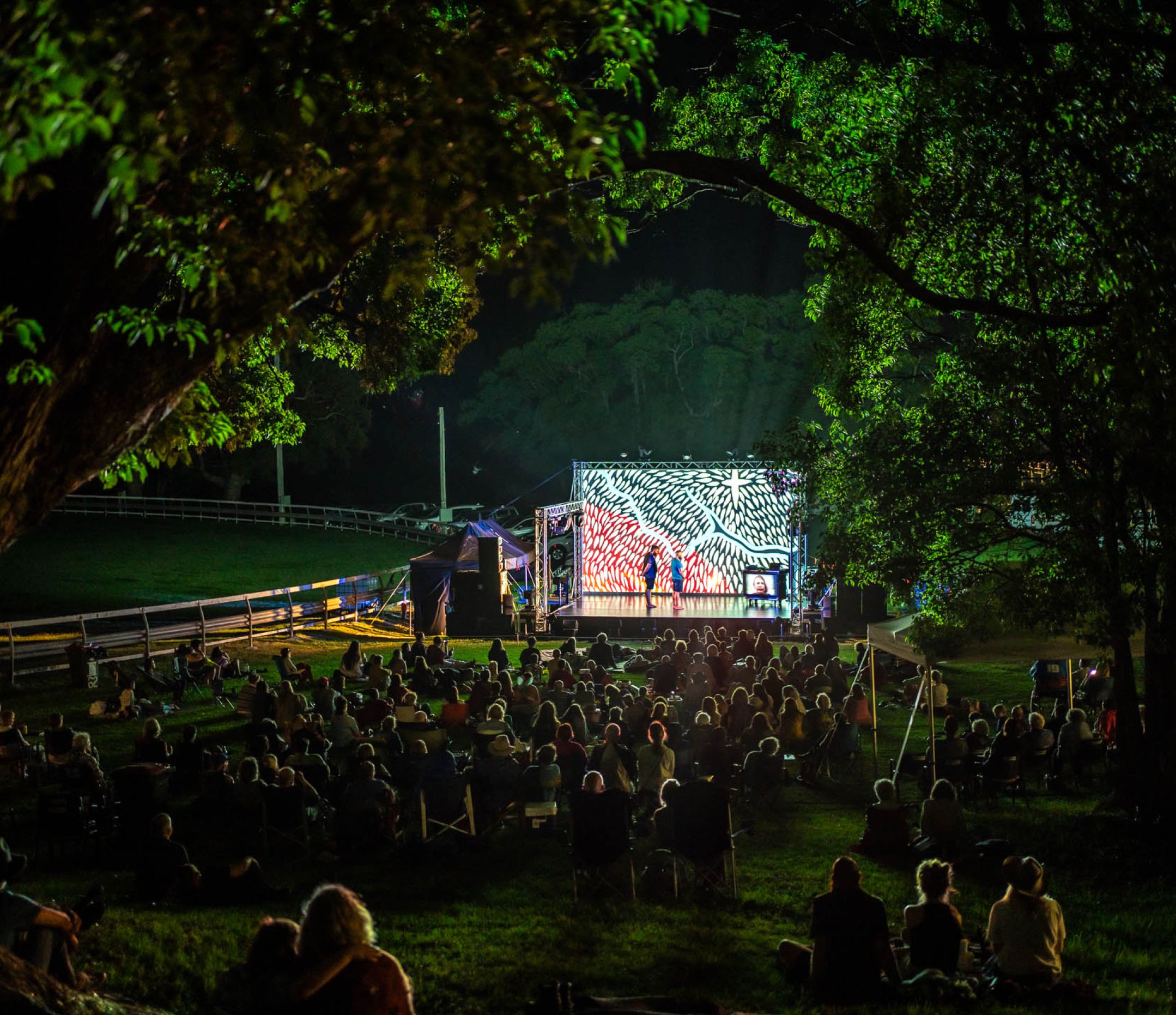 View of an outdoor night performance seen through a space in trees with the audience looking at a small stage with a projected image as the backdrop.