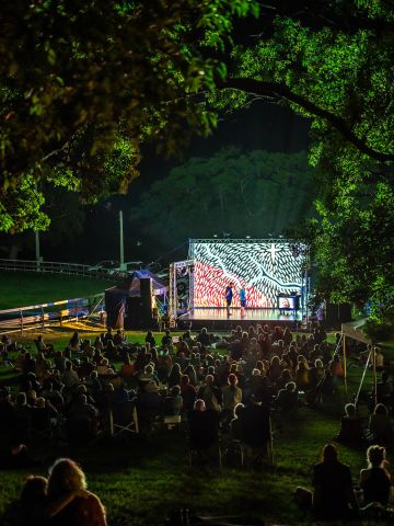 View of an outdoor night performance seen through a space in trees with the audience looking at a small stage with a projected image as the backdrop.