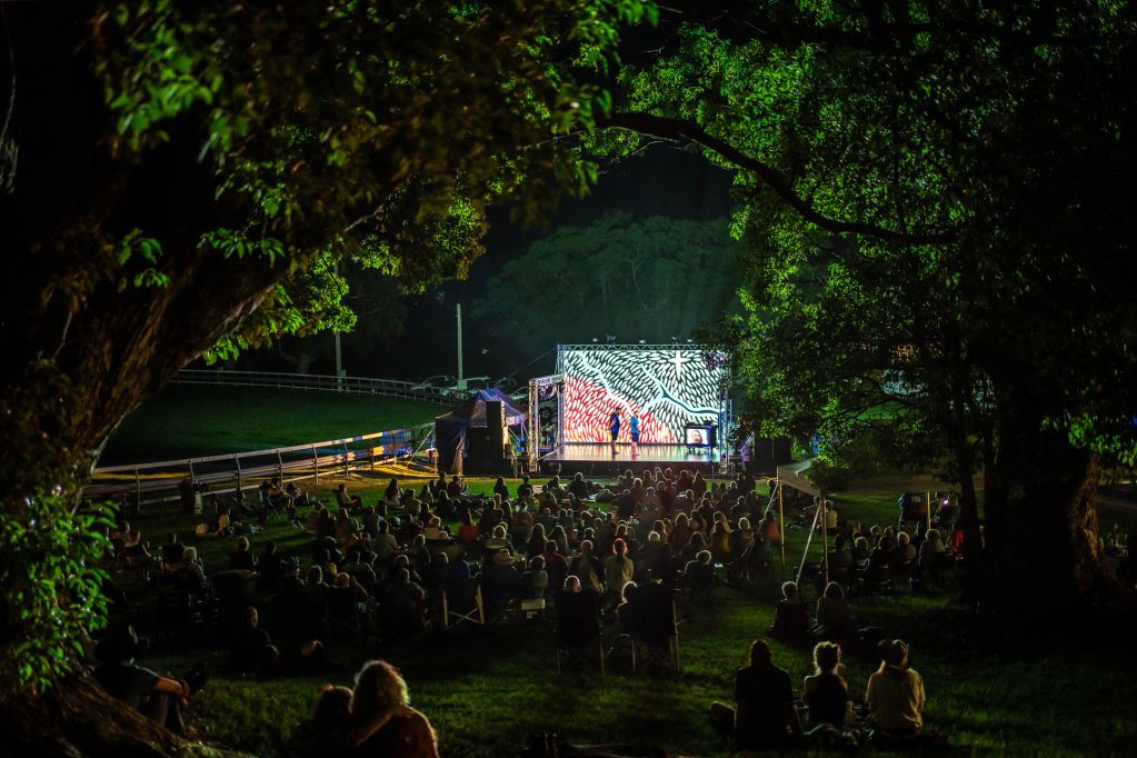 View of an outdoor night performance seen through a space in trees with the audience looking at a small stage with a projected image as the backdrop.