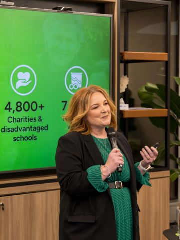 Middle-aged woman holding a microphone in front of a screen showing impact data (contained in post).