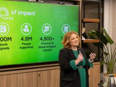 Middle-aged woman holding a microphone in front of a screen showing impact data (contained in post).