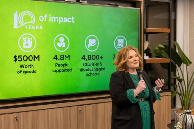 Middle-aged woman holding a microphone in front of a screen showing impact data (contained in post).