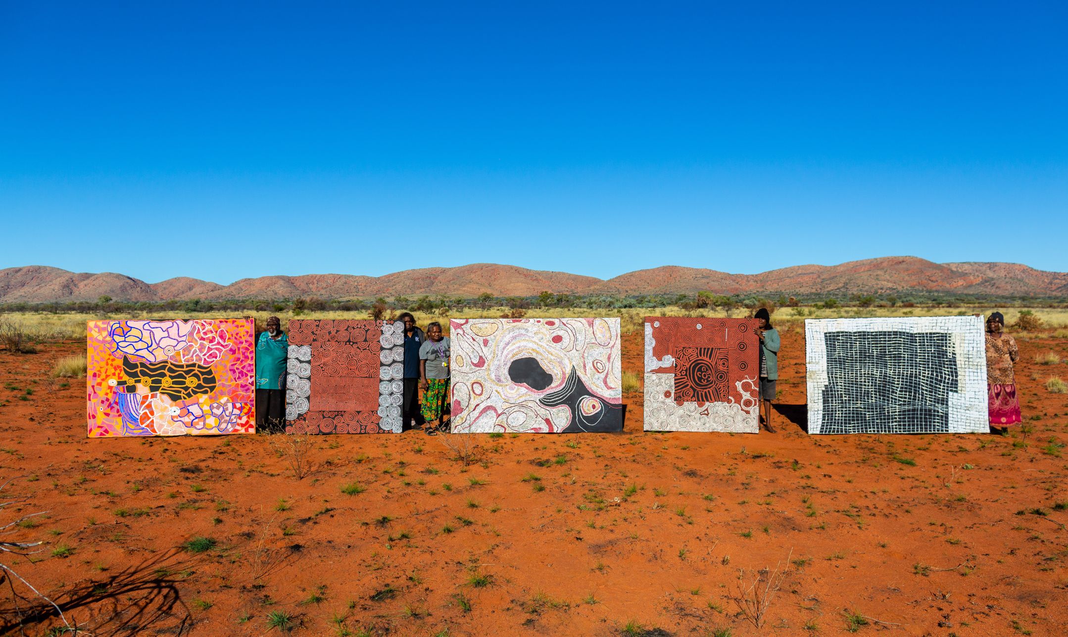 Indigenous artists standign next to theri large paintings in the desert. Red earth in the foreground, blue sky in the background.