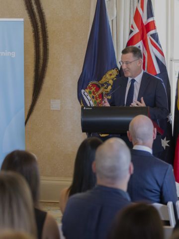 Middle-aged man in a suit standing at a lectern in front of Australian, Aboriginal and Torres Strair Island flags in front of a seated audience.