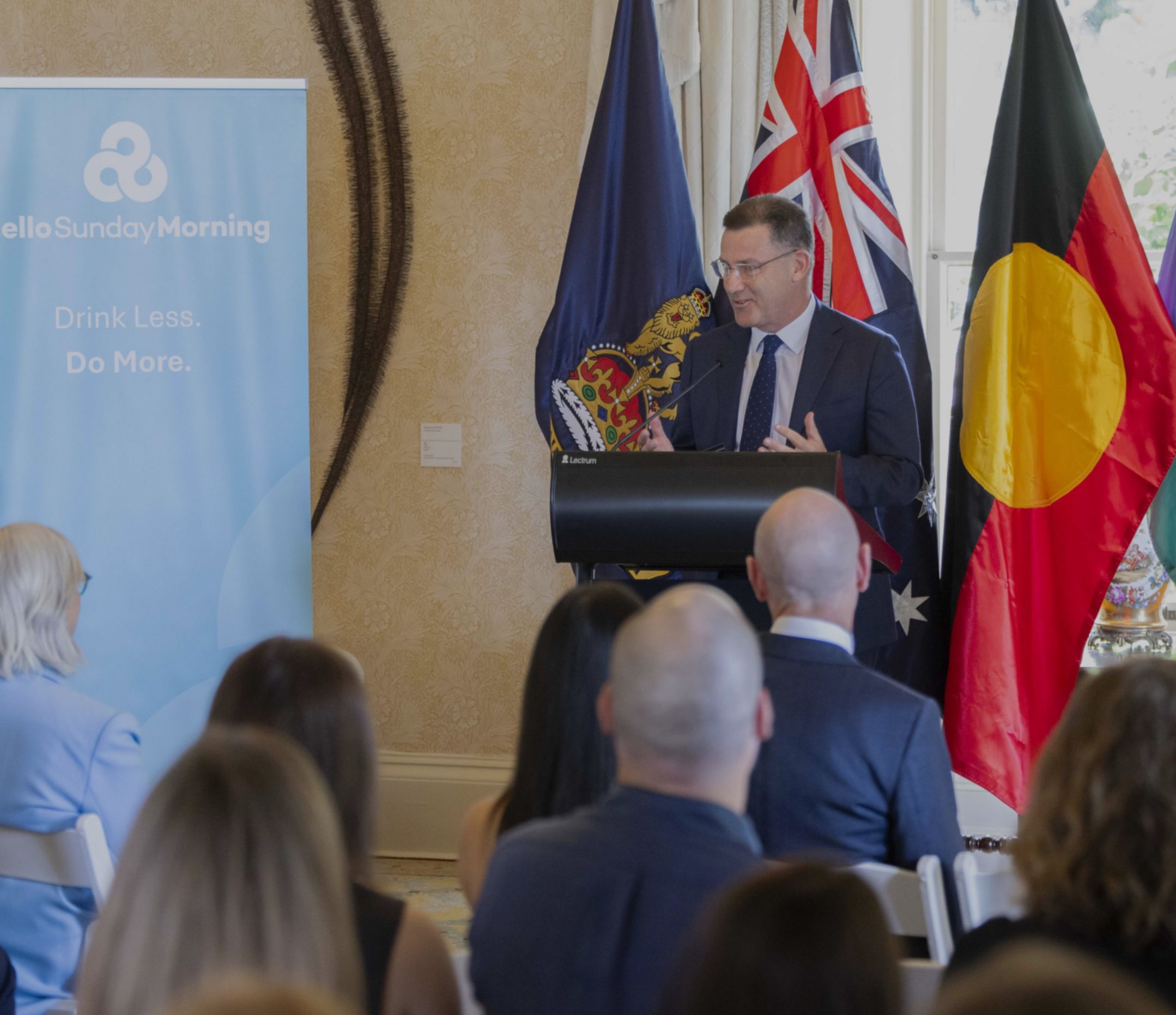 Middle-aged man in a suit standing at a lectern in front of Australian, Aboriginal and Torres Strair Island flags in front of a seated audience.