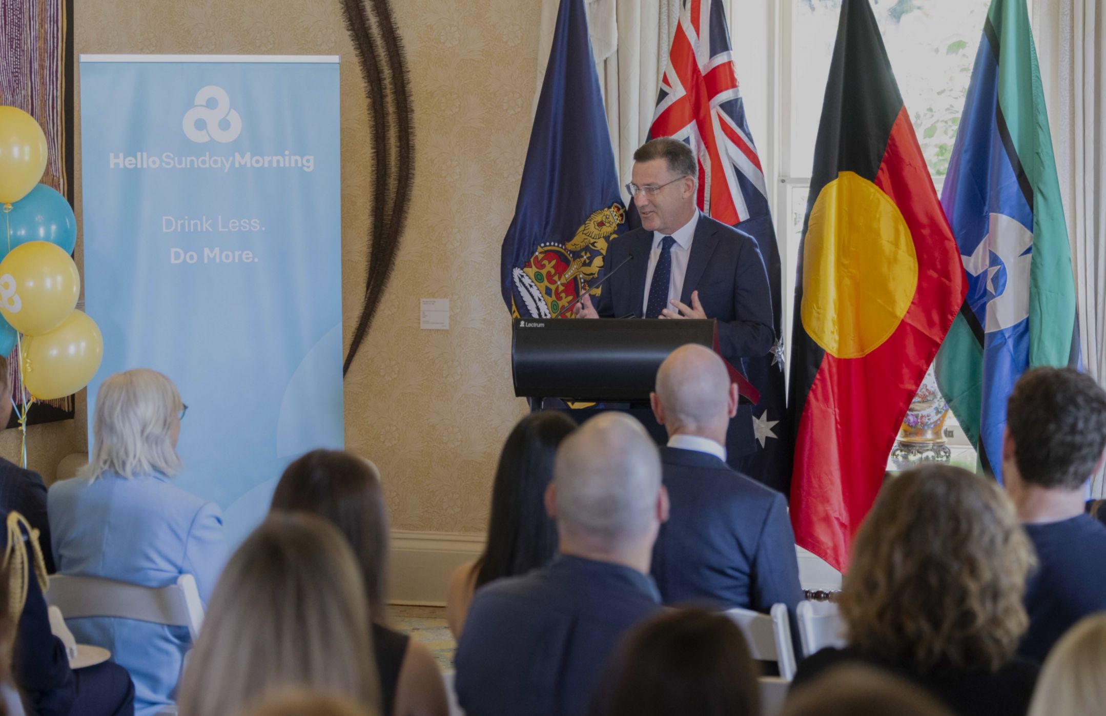 Middle-aged man in a suit standing at a lectern in front of Australian, Aboriginal and Torres Strair Island flags in front of a seated audience.