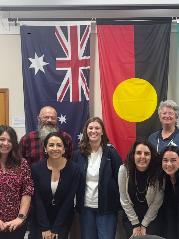 A group of 10 people smiling at the camera in front of the Australian, Aboriginal and Torres Strait Isalands flags.
