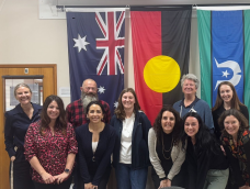 A group of 10 people smiling at the camera in front of the Australian, Aboriginal and Torres Strait Isalands flags.