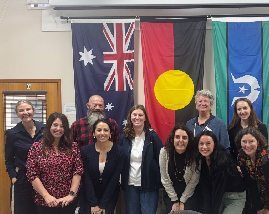 A group of 10 people smiling at the camera in front of the Australian, Aboriginal and Torres Strait Isalands flags.