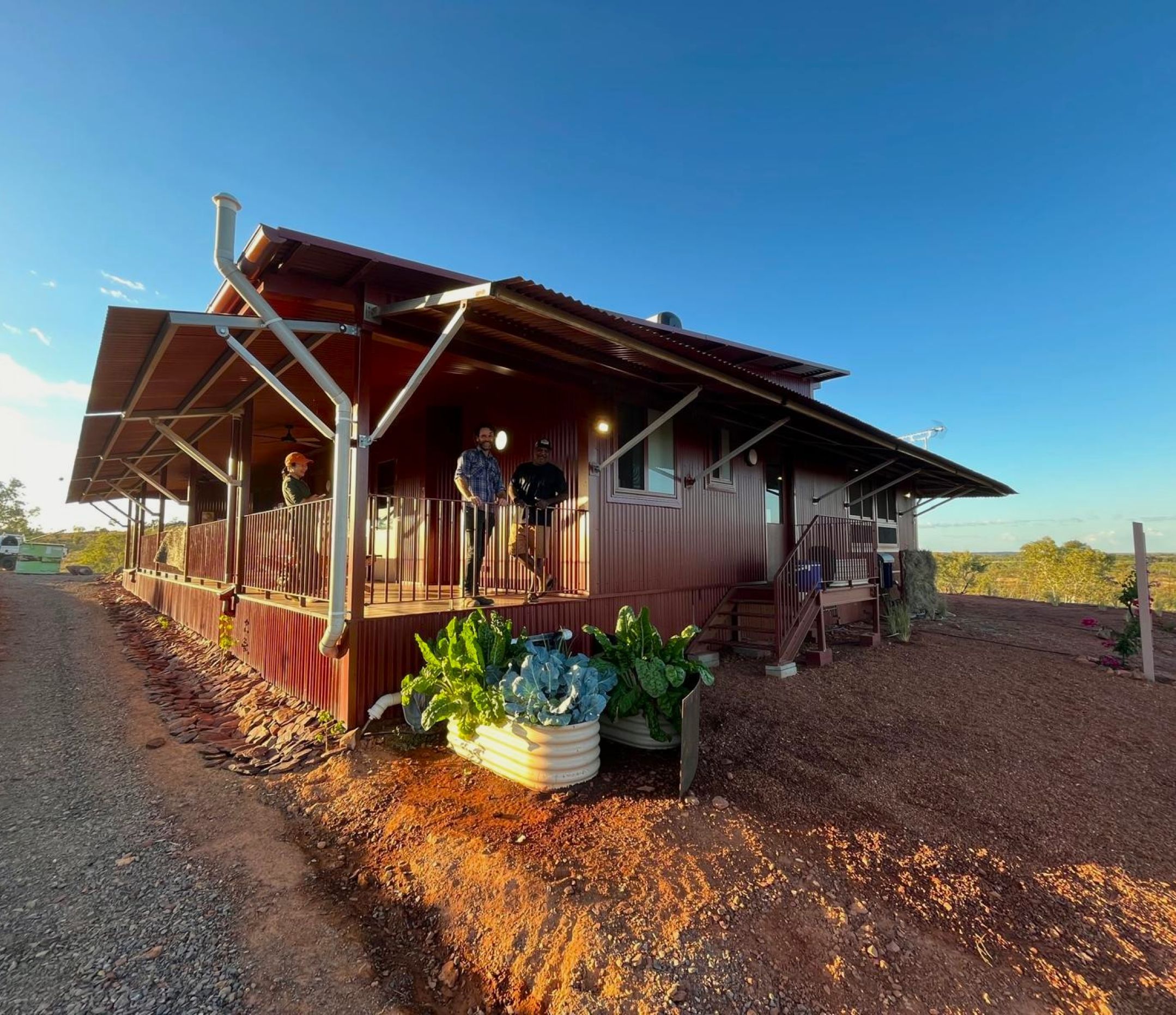 Two men standing on the verandah of a new house situated with red earth in the foregroudn and a blue sky in the background.