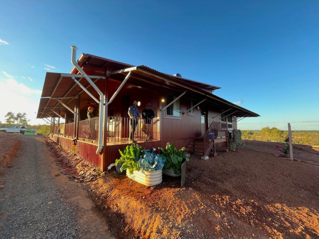 Two men standing on the verandah of a new house situated with red earth in the foregroudn and a blue sky in the background.