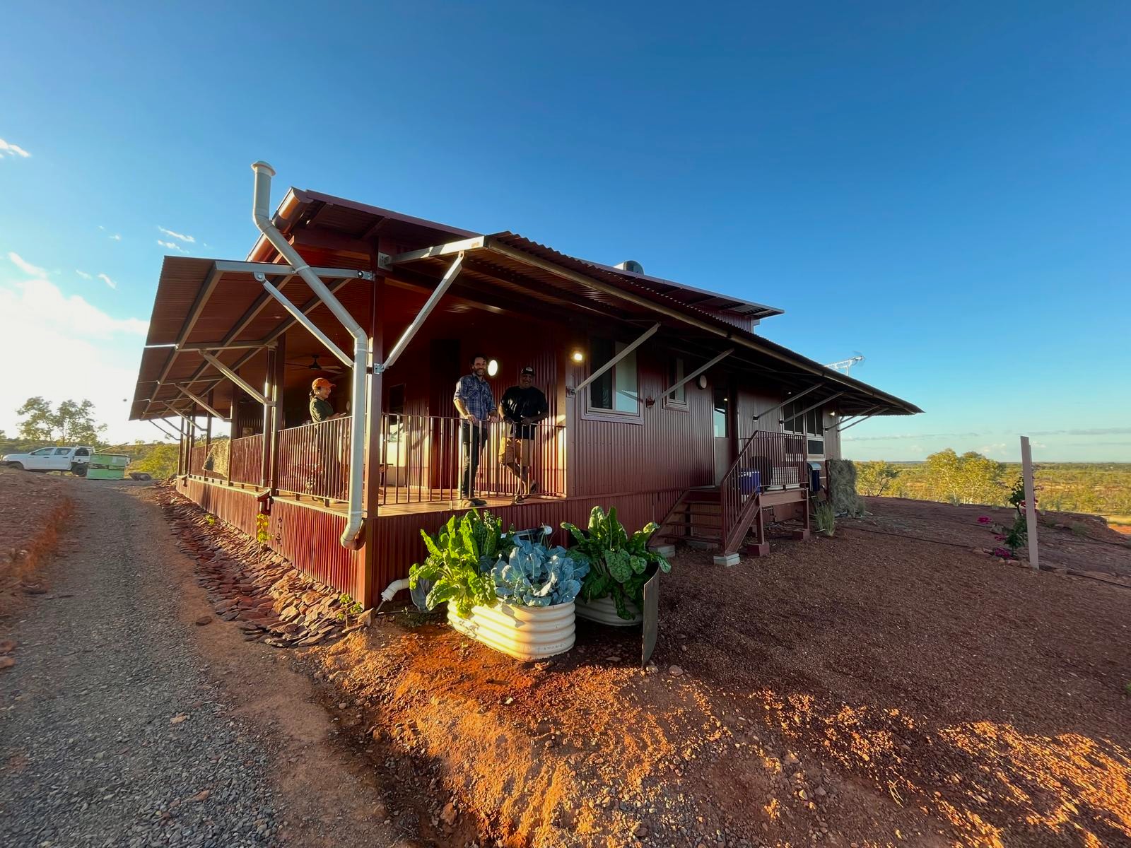 Two men standing on the verandah of a new house situated with red earth in the foregroudn and a blue sky in the background.