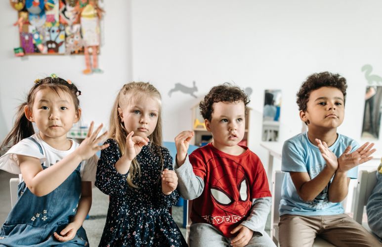 Four young children sitting next to each other gesturing with their hands