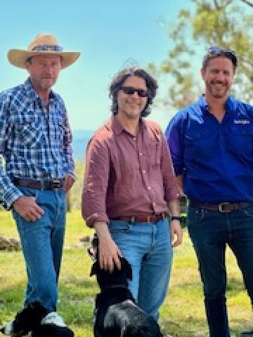 Four middle-aged men smiling, wearing jeans and check shirts standing in a paddock. One is patting a sheepdog. smil