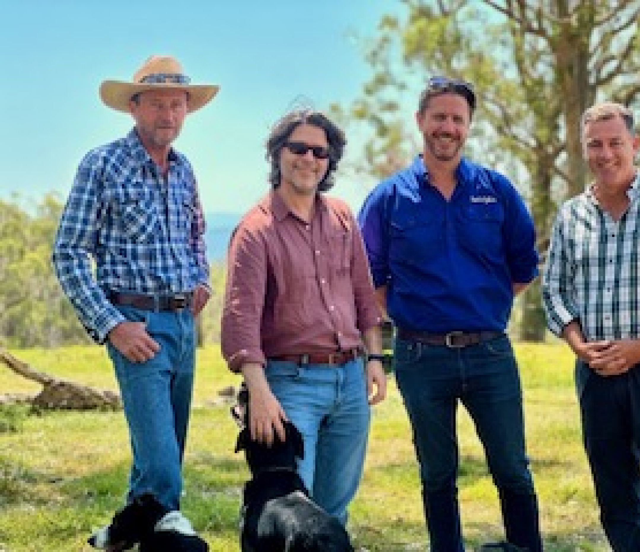 Four middle-aged men smiling, wearing jeans and check shirts standing in a paddock. One is patting a sheepdog. smil