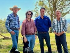 Four middle-aged men smiling, wearing jeans and check shirts standing in a paddock. One is patting a sheepdog. smil