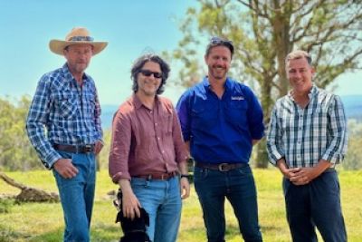 Four middle-aged men smiling, wearing jeans and check shirts standing in a paddock. One is patting a sheepdog. smil